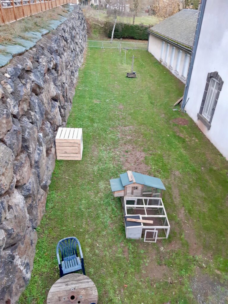 Composteur en bois entouré de verdure, près d'un petit chalet et de roches. Des plantes ornent les alentours. Idéal pour composter les déchets organiques des locataires de l'école.