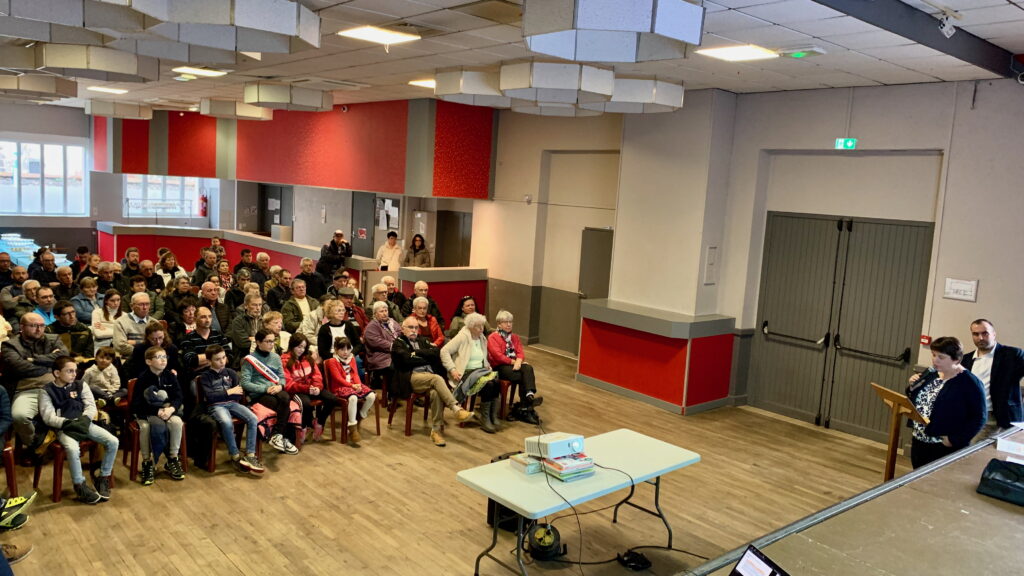 Vue intérieure de la salle des fêtes de Talizat lors de la cérémonie des vœux, avec une centaine de personnes assises face à une estrade où se trouve un pupitre et une table avec un vidéoprojecteur.